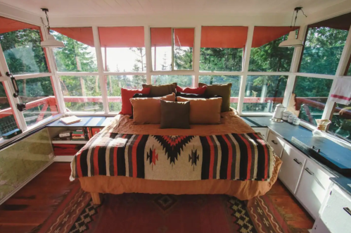 Cozy bedroom with a large bed, colorful blankets, and large windows overlooking a forested view.